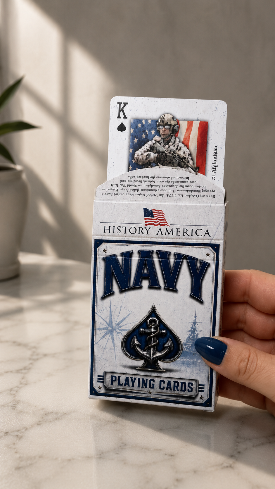 A hand with blue nail polish holds a box of History America Navy Playing Cards on a white marble surface with soft sunlight and shadows.