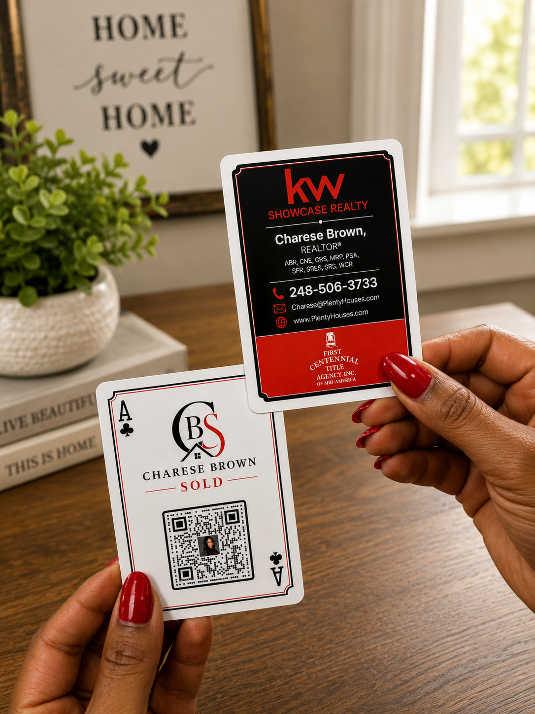 A person holds two playing cards in front of a window and framed wall art. One card is a black and red 'kw Showcase Realty' business card, and the other is an Ace of Spades playing card with 'Charese Brown Sold' and a QR code.
