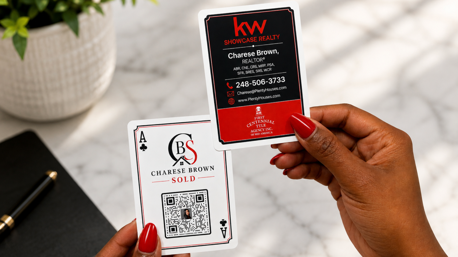 A person holds two realtor business cards in front of their face on a white marble surface. One card is black with red and white branding, and the other is white with a QR code and 'Charlese Brown Sold' text.