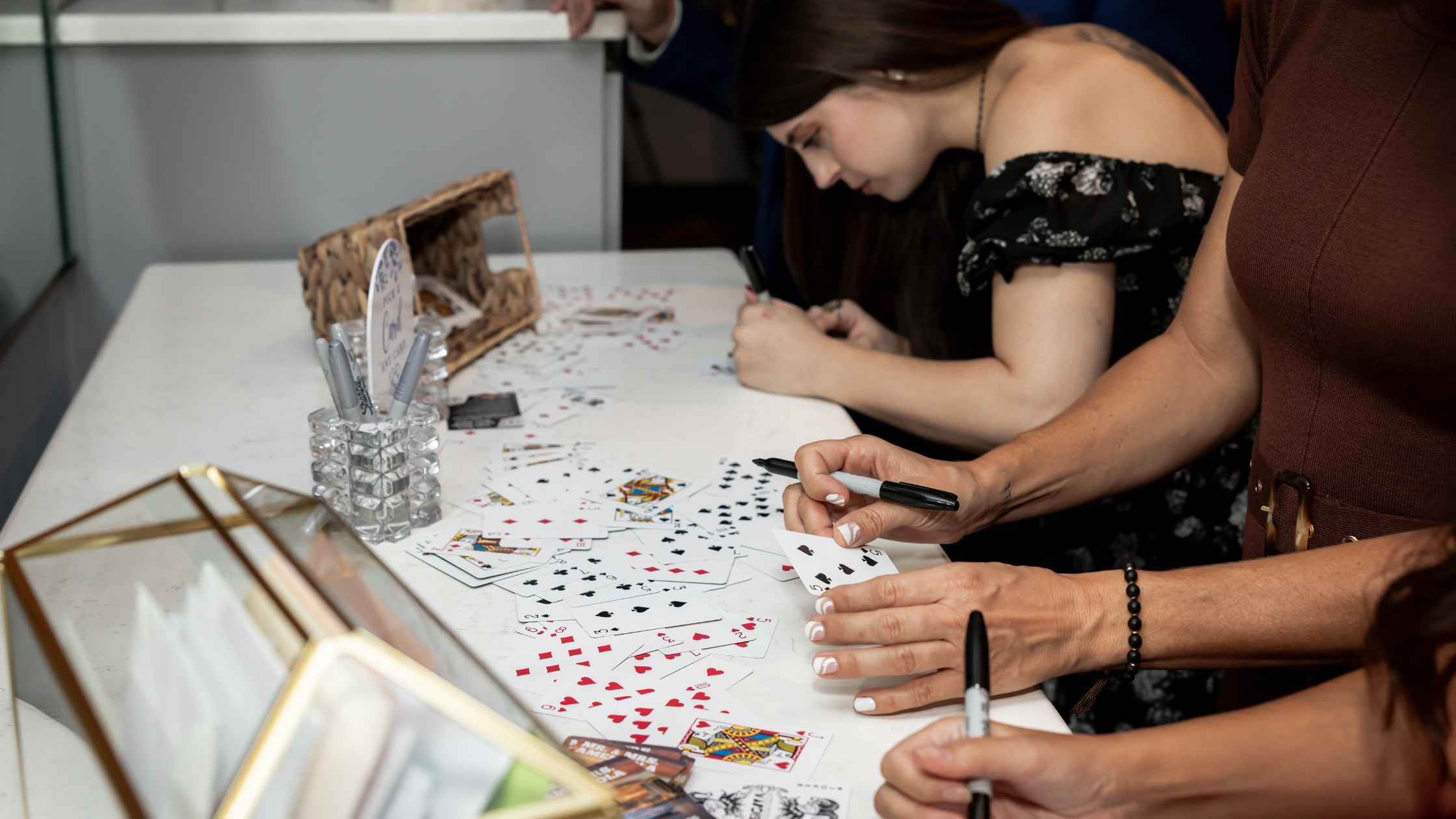 Several people gathered around a table playing with wedding playing cards. A woman in a black floral off-the-shoulder top is writing on a card, while others hold cards and pens.