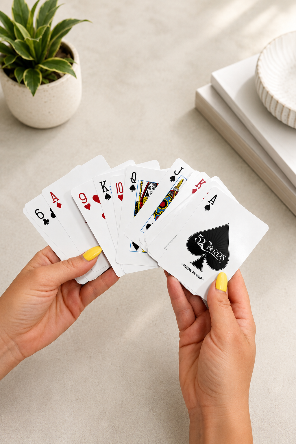 A person holds a fan of playing cards, including aces, kings, queens, and jacks, with a Queen of Spades card showing the back design. The hands have yellow nail polish, and the background is a light beige tabletop with a small potted succulent and stacked books.